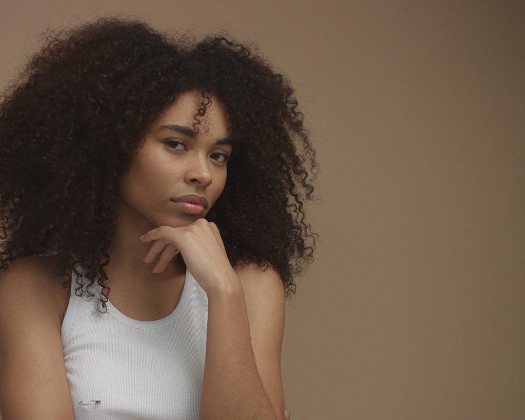 mixed race black woman portrait with big afro hair, curly hair in beige background. Thinking about something with a hand on chin